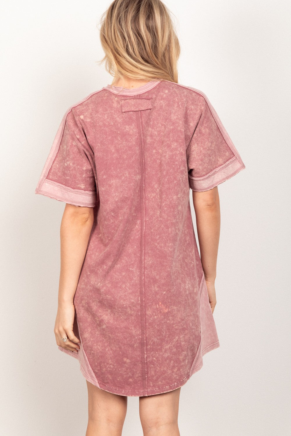 A woman stands against a plain background, wearing the loose-fitting, pink VERY J Short Sleeve V-Neck Tee Dress. Exuding casual style, she gazes at the camera with a slight smile while holding the side of her versatile piece.