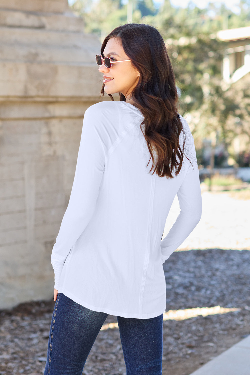 A woman wearing sunglasses and a slightly stretchy Basic Bae Full Size Round Neck Long Sleeve T-Shirt in white stands on a sidewalk with an arched structure in the background, smiling slightly.