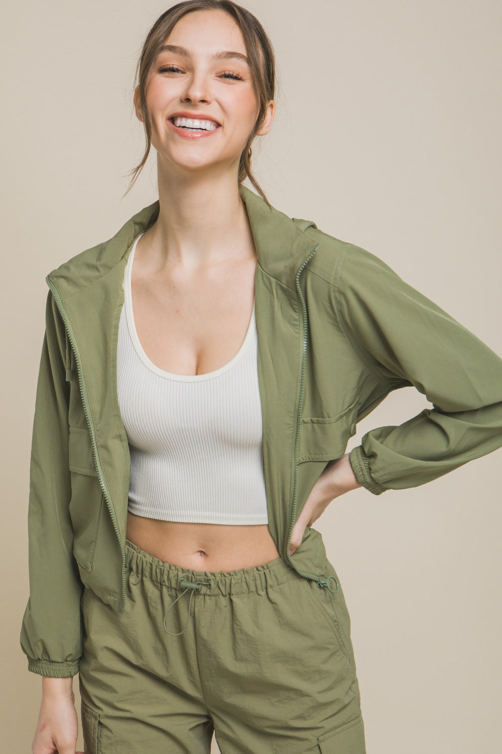 A smiling person is wearing a Love Tree Drawstring Hem Hooded Zip-Up Jacket in light green with matching pants, complemented by a white top underneath, and is standing against a plain background.