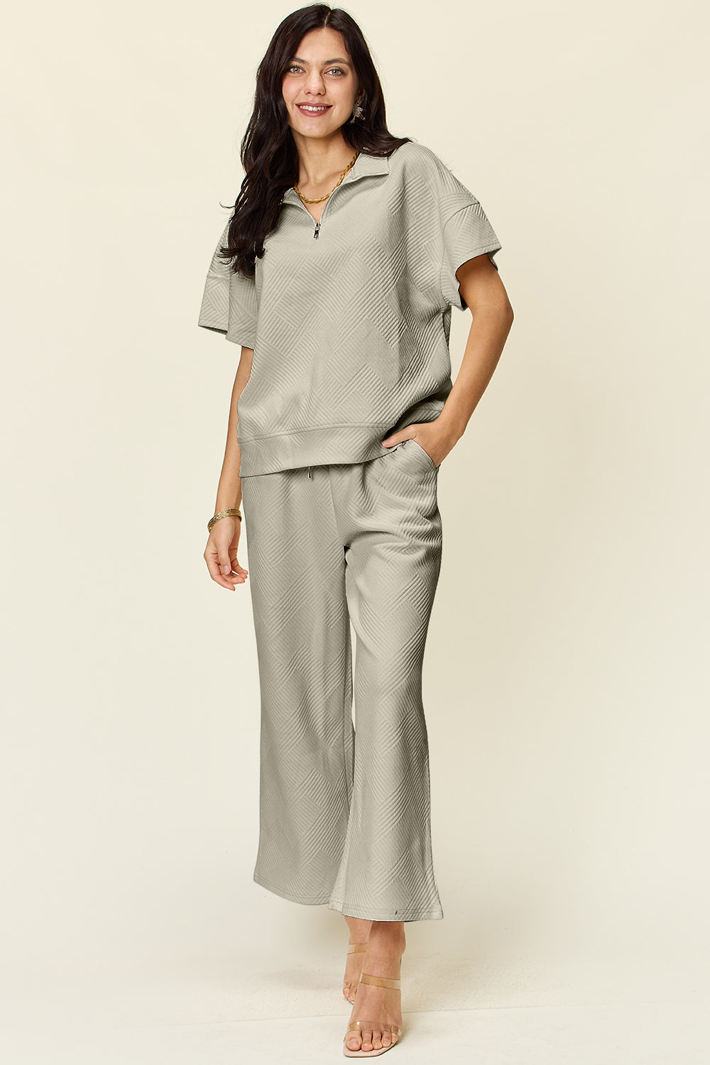 A woman is wearing the Double Take Full Size Texture Half Zip Short Sleeve Top and Pants Set, featuring a white zip-up short-sleeve top and matching wide-leg, drawstring pants. She stands with one hand in her pocket and smiles against a light background.