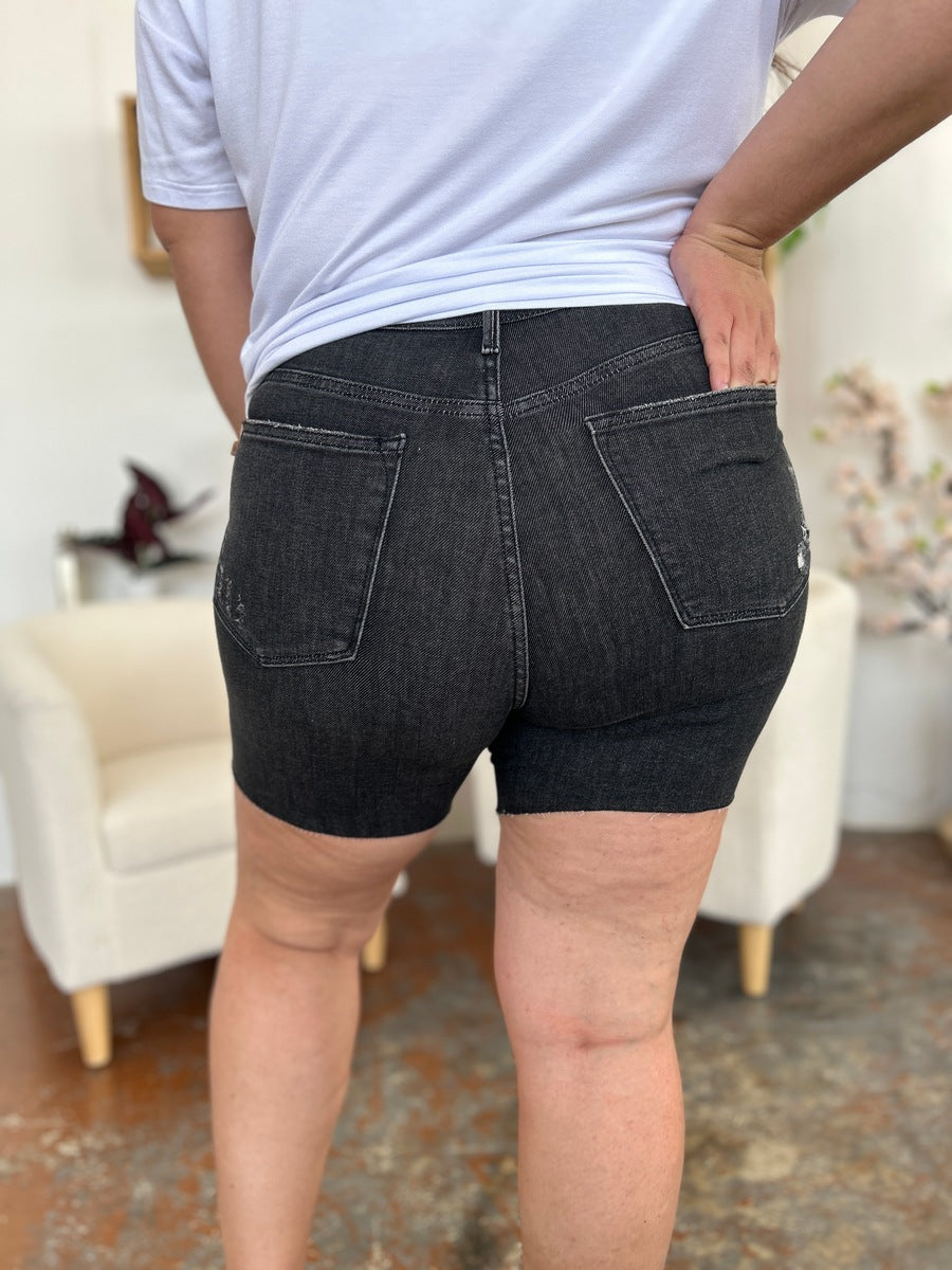 A person wearing a white t-shirt and Judy Blue Full Size High Waist Rigid Magic Denim Shorts is standing indoors in front of a beige chair.