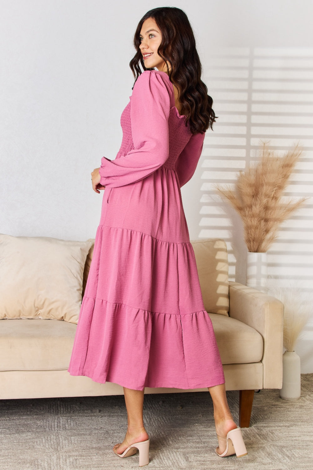 A woman in a Culture Code Full Size Ruffle Trim Smocked Tiered Dress stands in front of a beige sofa adorned with decorative pillows and framed by dried pampas grass, embodying a graceful silhouette perfect for elegant evening affairs.