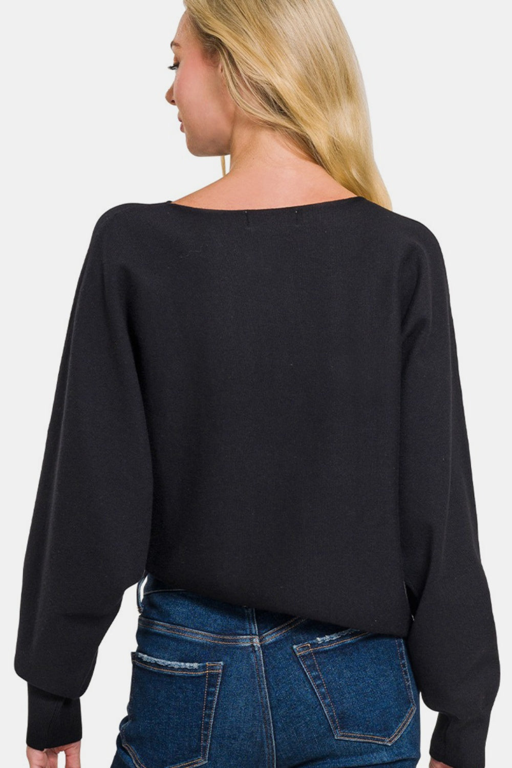 A woman with long blonde hair is wearing a Zenana Asymmetric Hem Long Sleeve Sweater in black paired with blue jeans, standing against a plain white background.