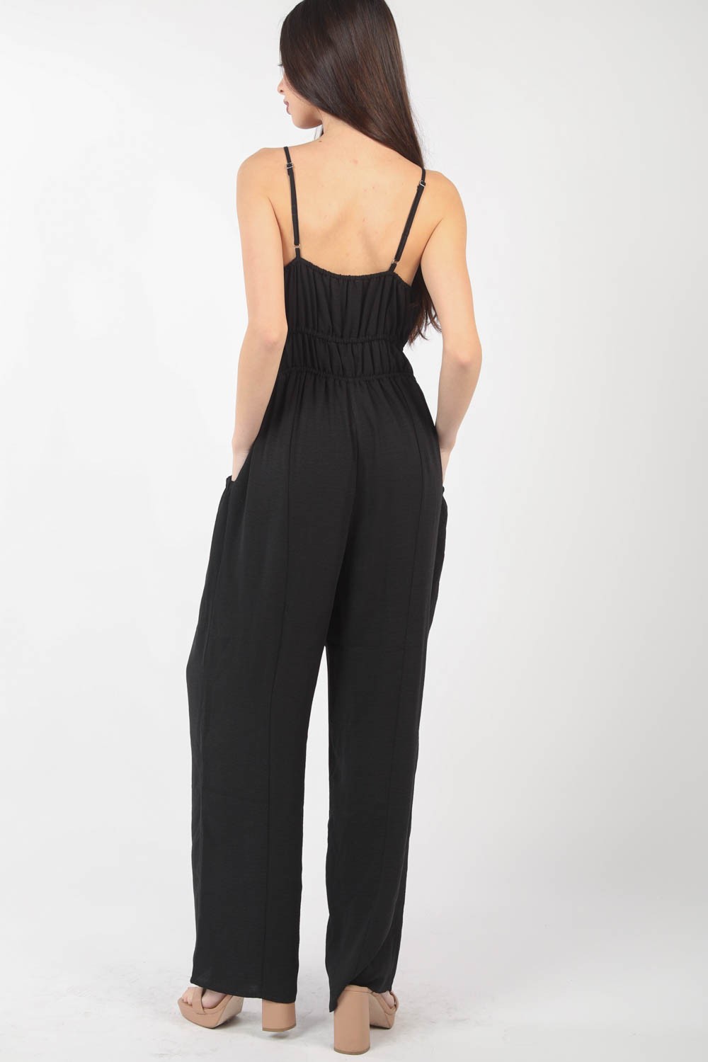 A woman stands against a plain backdrop wearing the VERY J Pintuck Detail Woven Sleeveless Jumpsuit. The black jumpsuit has thin straps, wide-leg pants, and is crafted from woven fabric with subtle pintuck details. She has her hands in her pockets and is looking at the camera.