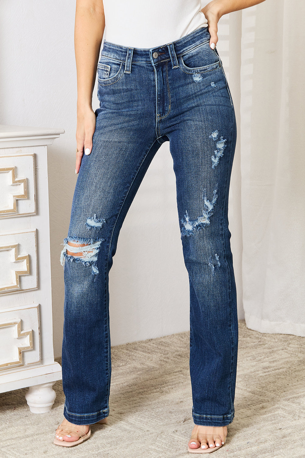 A person wearing Judy Blue Full Size Mid Rise Hand Sand & Destroy Bootcut Jeans stands with hands in pockets. The slightly stretchy jeans have rips on both legs, and the person is standing indoors beside a white dresser.