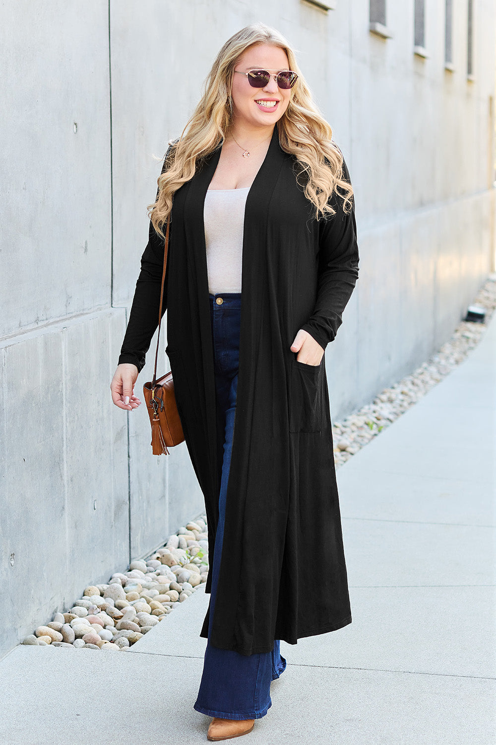 A woman stands against a concrete wall, wearing the Basic Bae Full Size Open Front Long Sleeve Cover Up made from rayon spandex, a white top, blue jeans in her perfect size, and brown shoes with her hands in her pockets. Machine wash cold for best results.