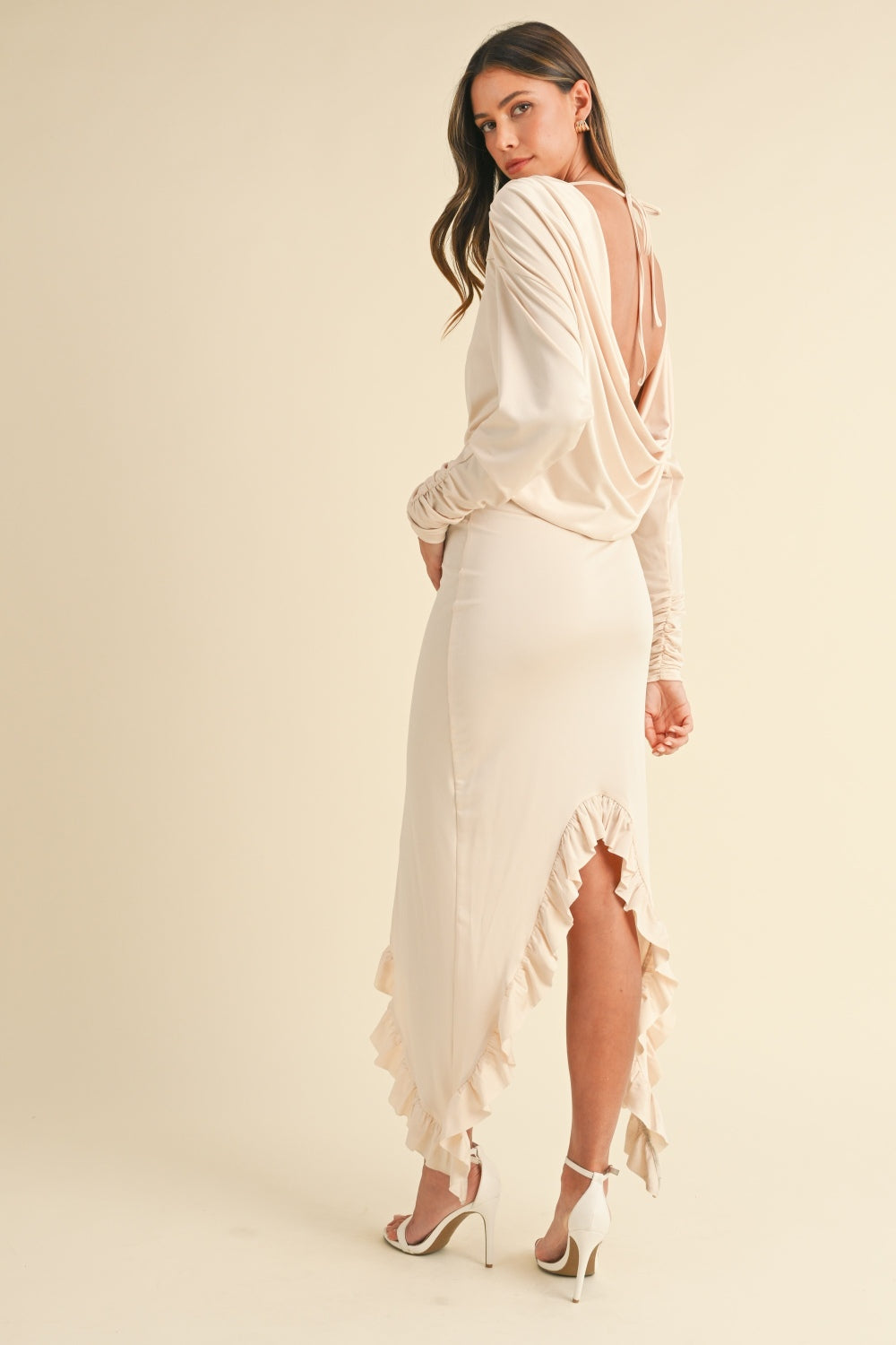 A woman stands in the Mable Backless Asymmetric Ruffle Hem Dress paired with white high-heeled sandals against a plain background.