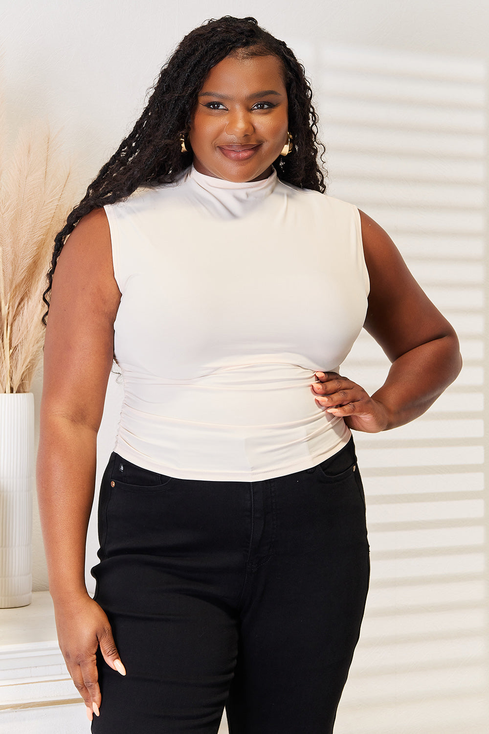 A person wearing a sleeveless white top and black pants stands indoors, with their hand resting on their pants' pocket and light streaming through blinds in the background. The Mock Neck Ruched Cropped Tank adds a touch of summer style to the scene.