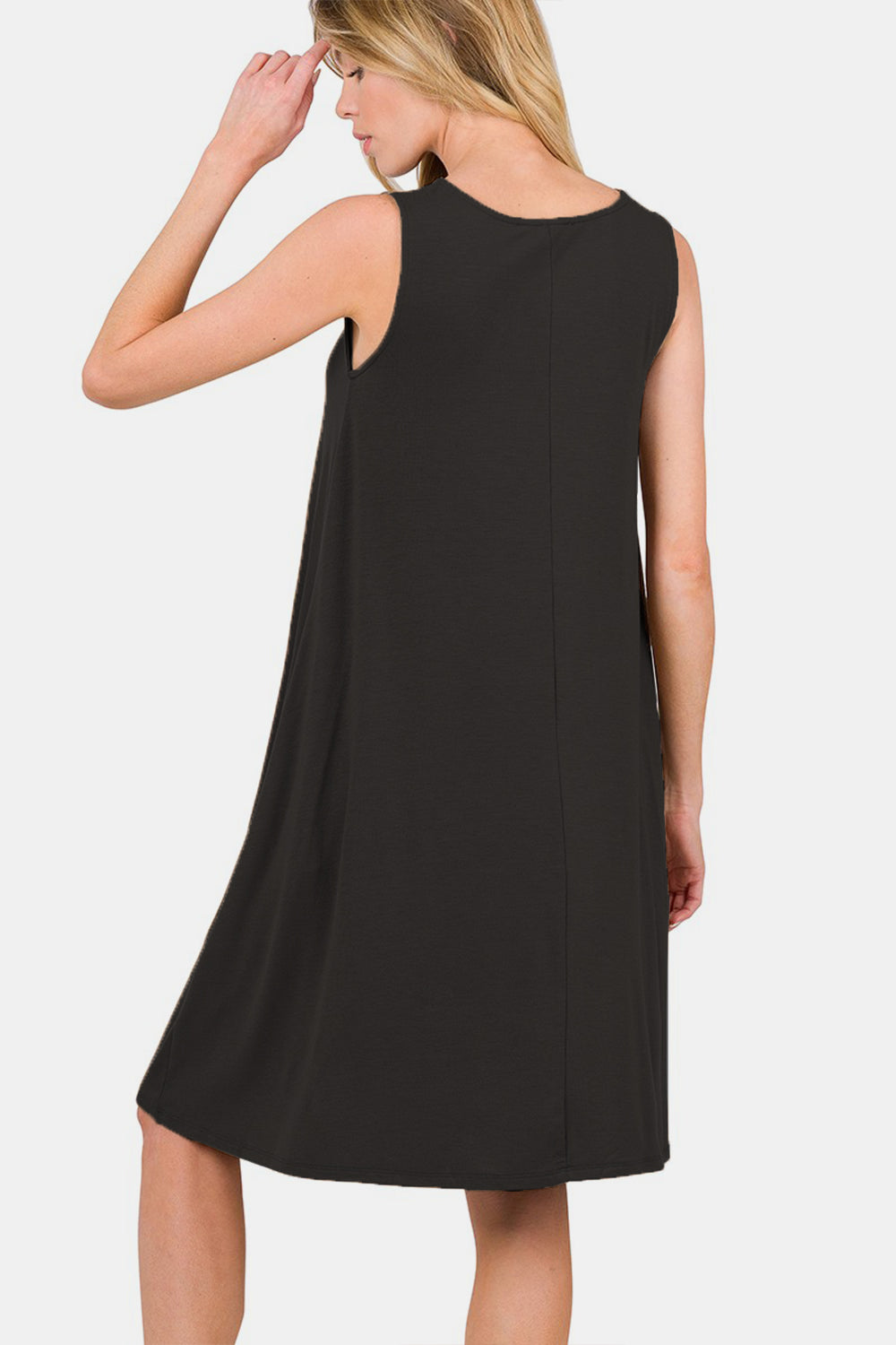 A woman with blonde hair smiles while wearing the Zenana Full Size Sleeveless Flared Dress with Side Pockets in black. She holds one side of the dress with her hand and has her other hand in the pocket, showcasing this versatile wardrobe piece.