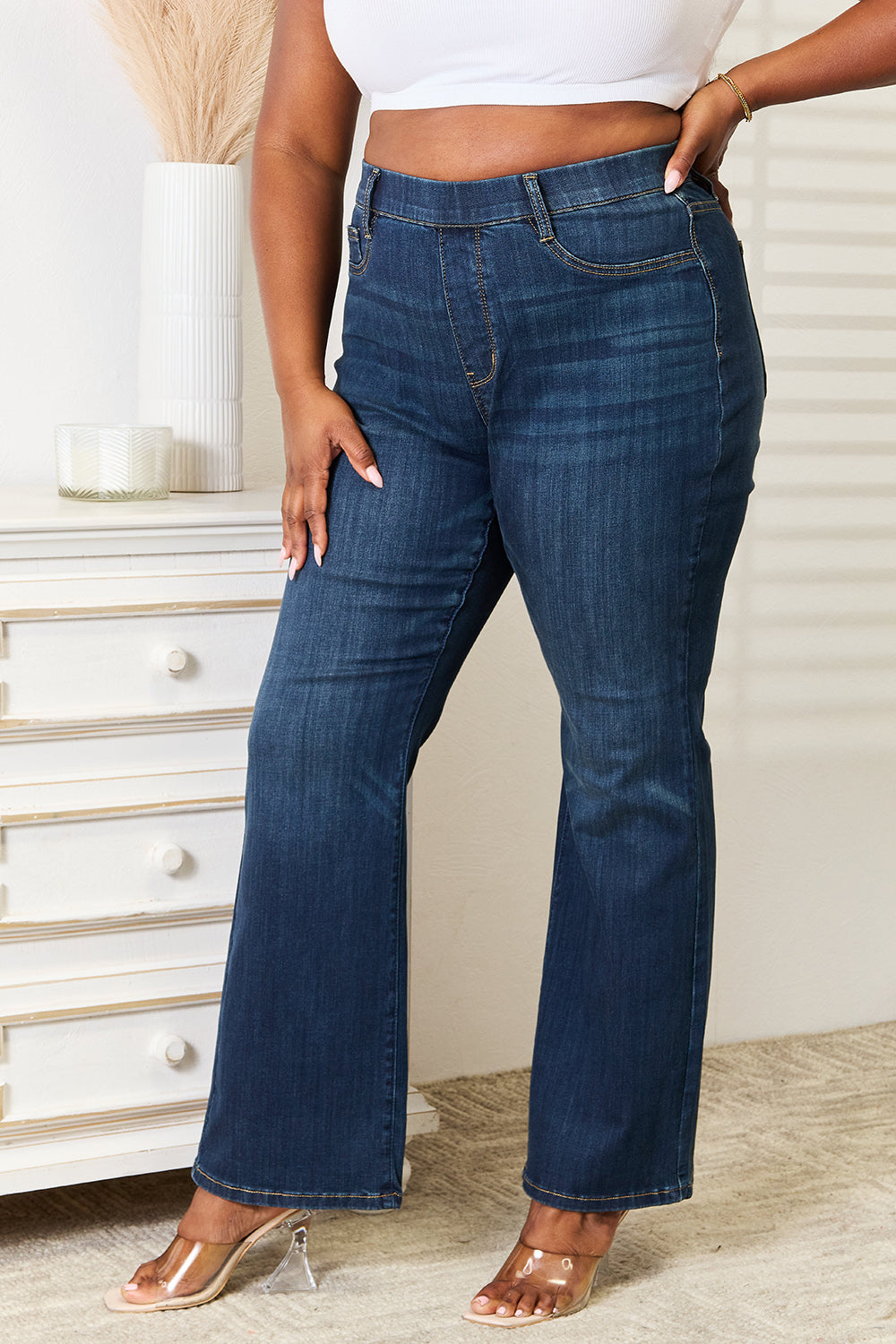 A person wearing a white crop top and Judy Blue Full Size Elastic Waistband Slim Bootcut Jeans stands next to a white dresser.