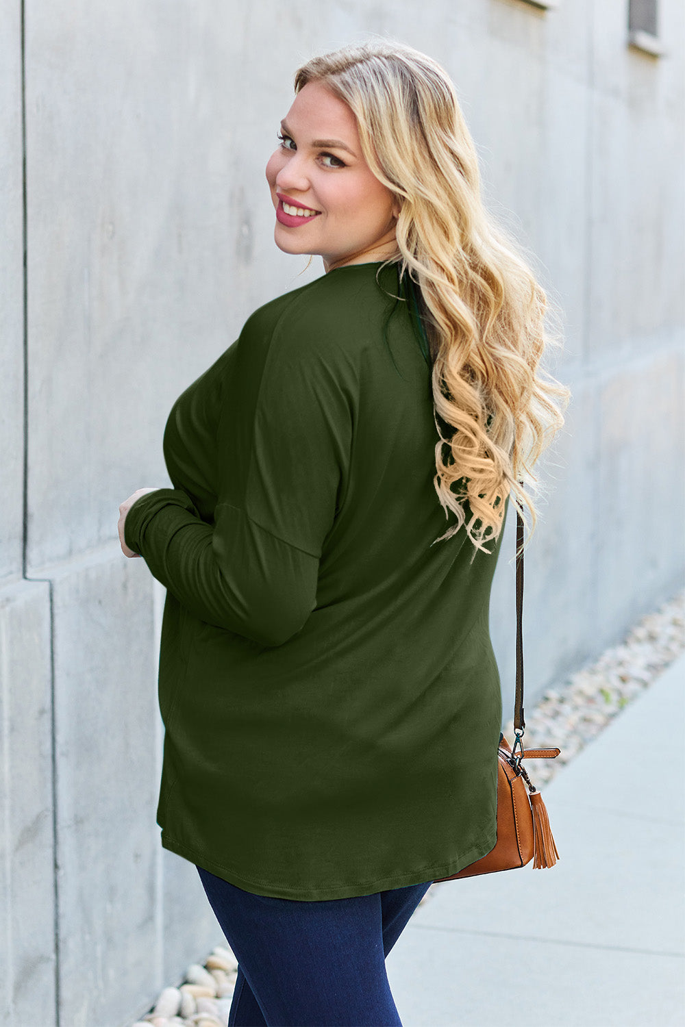 A woman with shoulder-length hair wearing a Basic Bae Full Size Round Neck Dropped Shoulder T-Shirt in green and a pair of jeans, exuding a basic style, stands against a concrete wall. She's holding an imported brown woven backpack on her shoulder while touching her hair with her hand.