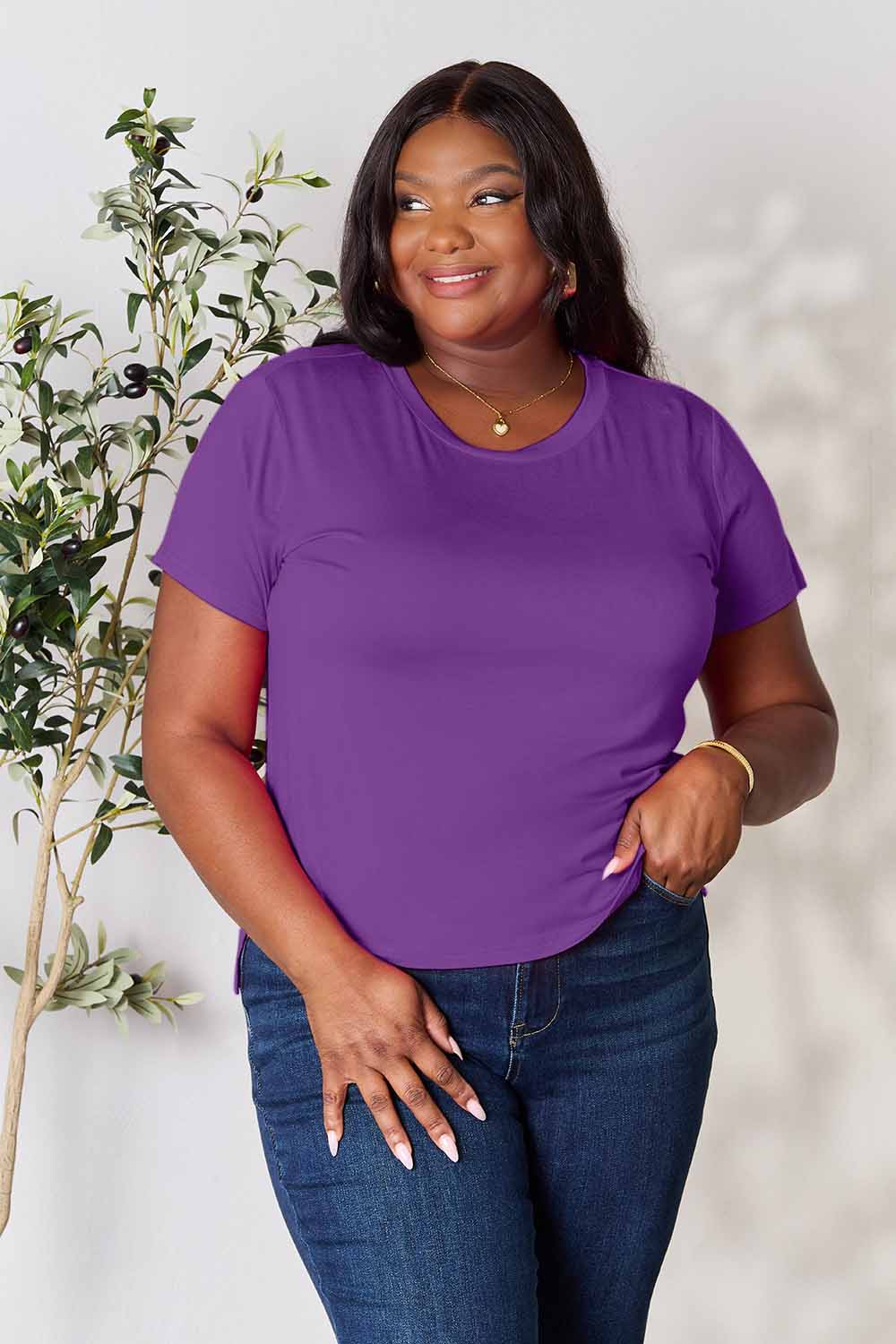 A woman stands next to an indoor plant, smiling, with one hand in her khaki pants pocket. In her pink Basic Bae Full Size Round Neck Short Sleeve T-Shirt and gold jewelry, she radiates casual elegance.