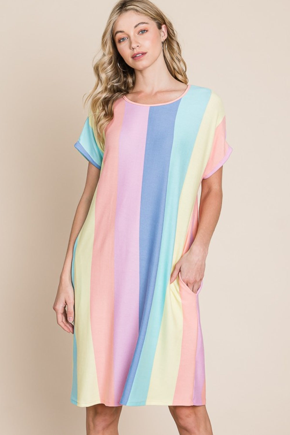 A person stands against a plain background, wearing the BOMBOM Striped Short Sleeve Dress with Pockets, crafted from comfortable fabric and featuring pastel stripes, with hands casually tucked into the convenient pockets.