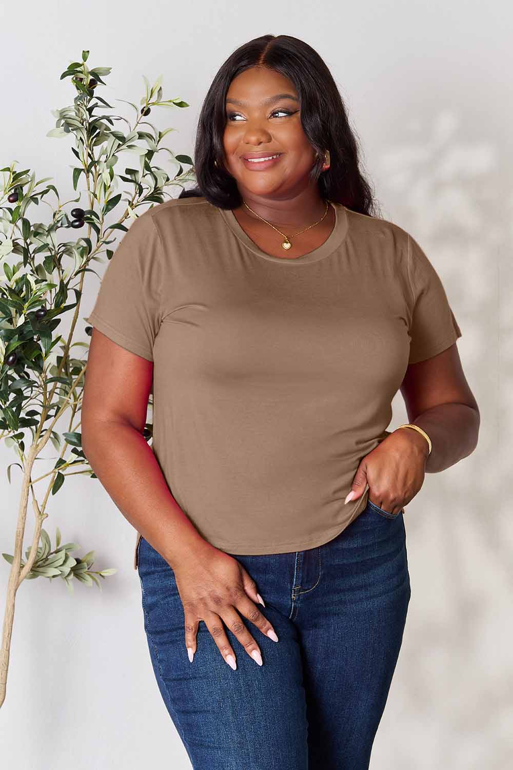 A woman stands next to an indoor plant, smiling, with one hand in her khaki pants pocket. In her pink Basic Bae Full Size Round Neck Short Sleeve T-Shirt and gold jewelry, she radiates casual elegance.