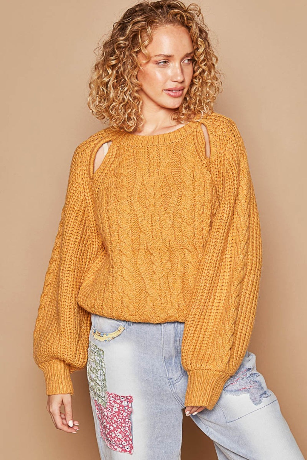 A person with curly hair wears a POL Cable Knit Cutout Long Sleeve Sweater in mustard, paired with light blue jeans adorned with patchwork designs.