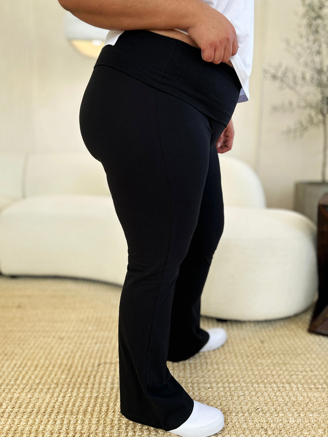 An individual dressed in Zenana Full Size High Waist Wide Waistband Bootcut Active Pants and white shoes, standing with a hand on the hip in front of a setting that includes a white couch and a plant.