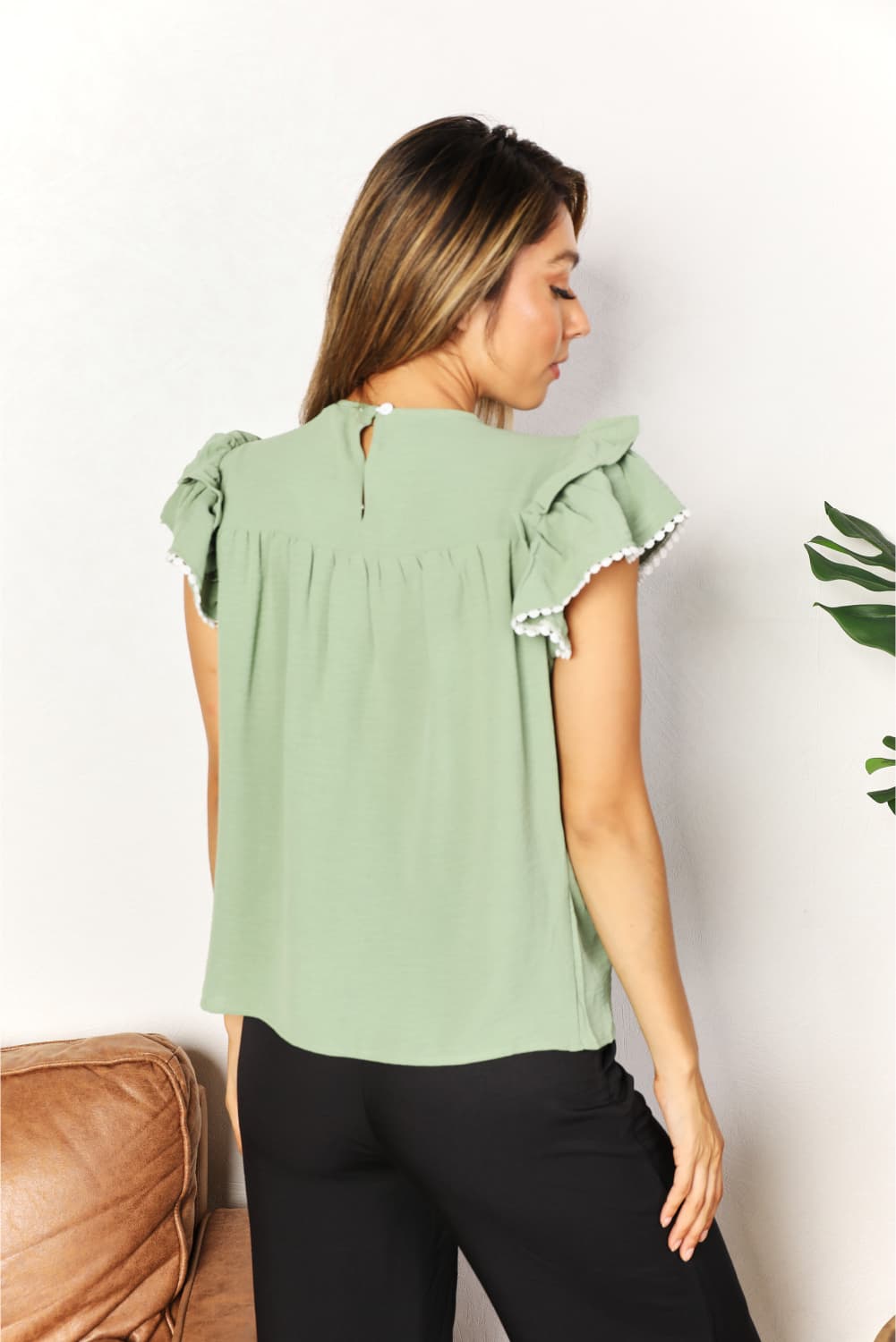 A woman stands and smiles, wearing the Perfee Pleated Detail Flutter Sleeve Blouse in light green paired with black pants.