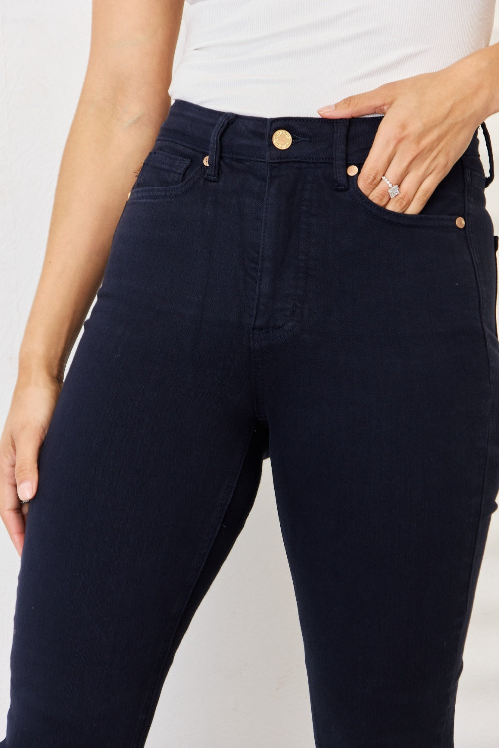 A person wearing a white sleeveless top, Judy Blue Full Size Garment Dyed Tummy Control Skinny Jeans in dark blue, and light-colored slip-on shoes stands indoors with hands adjusting the jeans waistband. Shadows of horizontal lines are visible on the wall.