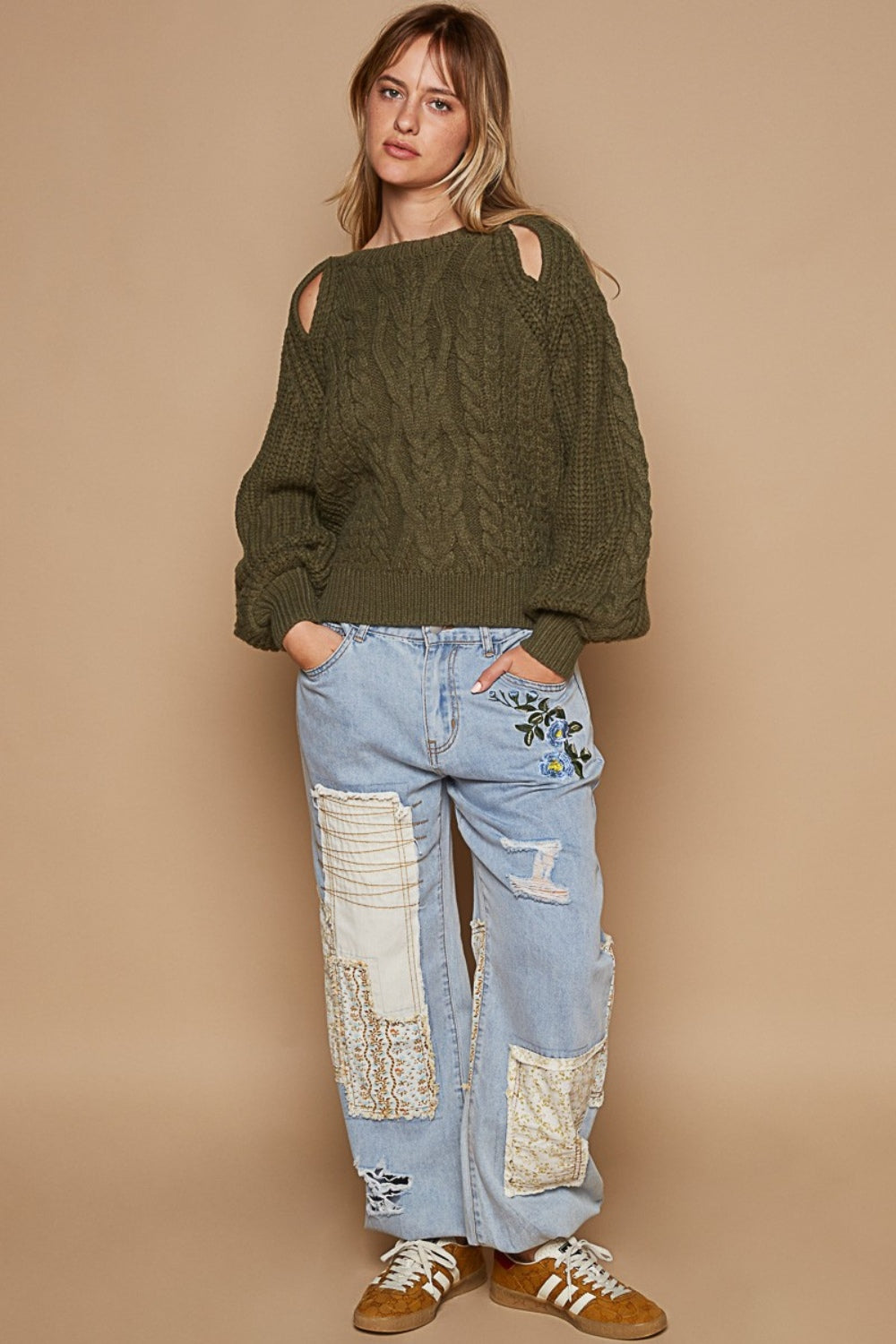 A woman wears the POL Cable Knit Cutout Long Sleeve Sweater in olive-green, showcasing a subtle shoulder keyhole, beautifully matched with light blue jeans adorned with floral embroidery, all set against a beige background.