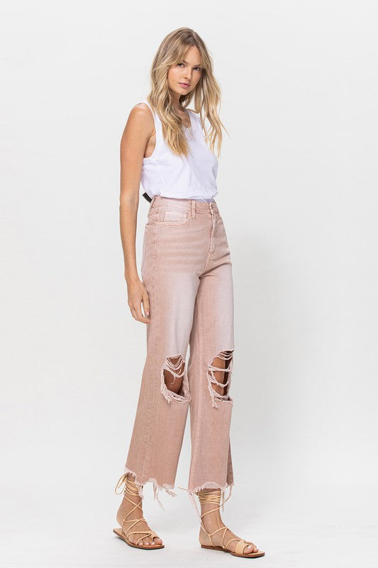 Person wearing 90's Vintage Crop Flare Jeans in rose pink with high waist, distressed details, rips on the knees, and frayed hems, paired with beige strappy sandals.