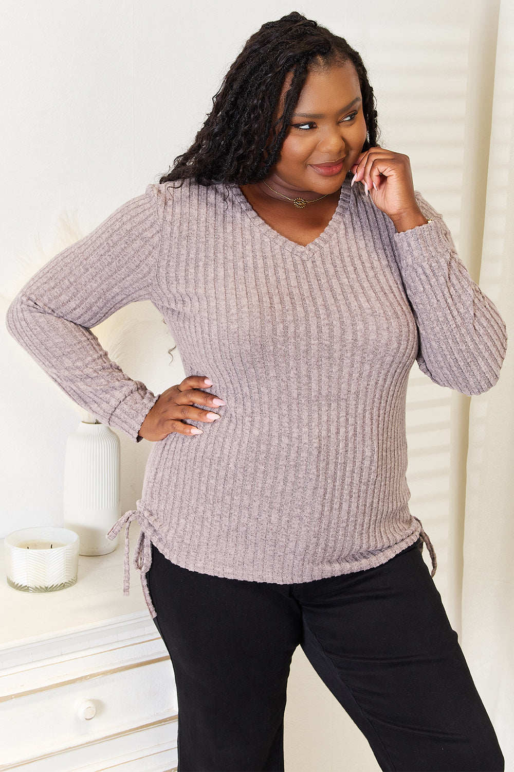 A woman stands indoors against a neutral background, wearing the "Double Take Drawstring Ribbed Long Sleeve T-Shirt" in light purple paired with dark pants. She is smiling and posing with one hand on her hip.