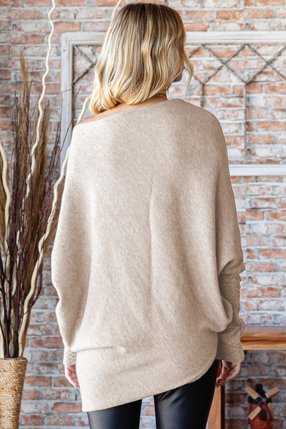 A woman with blonde hair wears a First Love Full Size Asymmetrical Hem Dolman Sleeve Sweater in beige and black leggings, standing against a brick wall background.