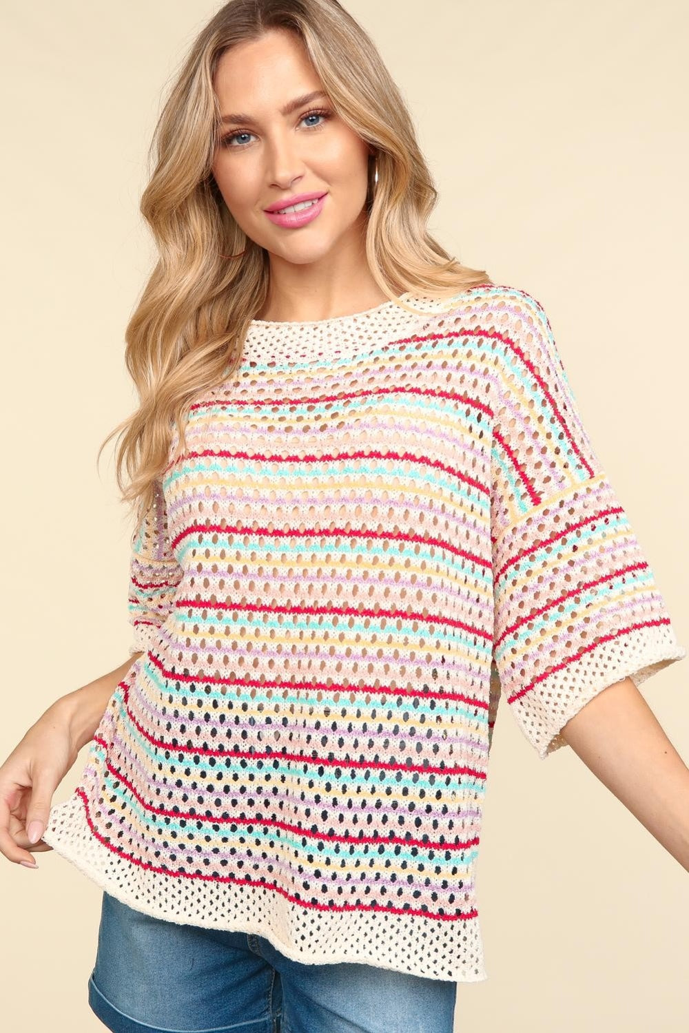 A woman stands smiling against a beige background, wearing the Haptics Striped Crochet Drop Shoulder Knit Top paired with denim shorts.