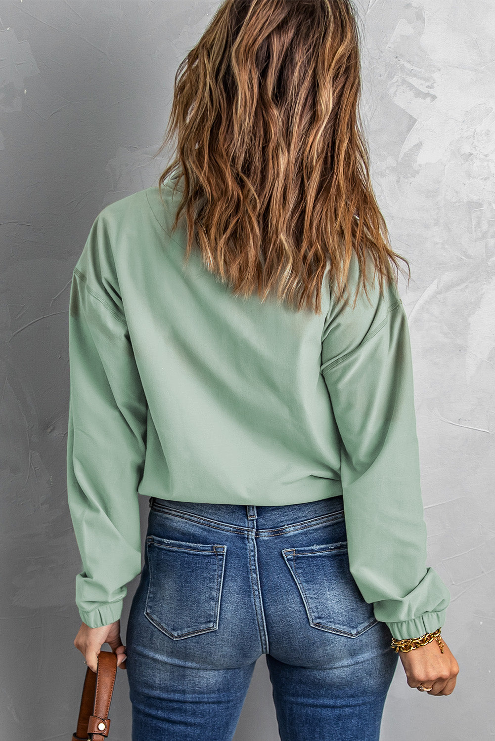 A person with wavy hair stands with their back to the camera, wearing a Green Zip Front Pocketed Pullover Sweatshirt and blue jeans, holding a small brown bag in their left hand.