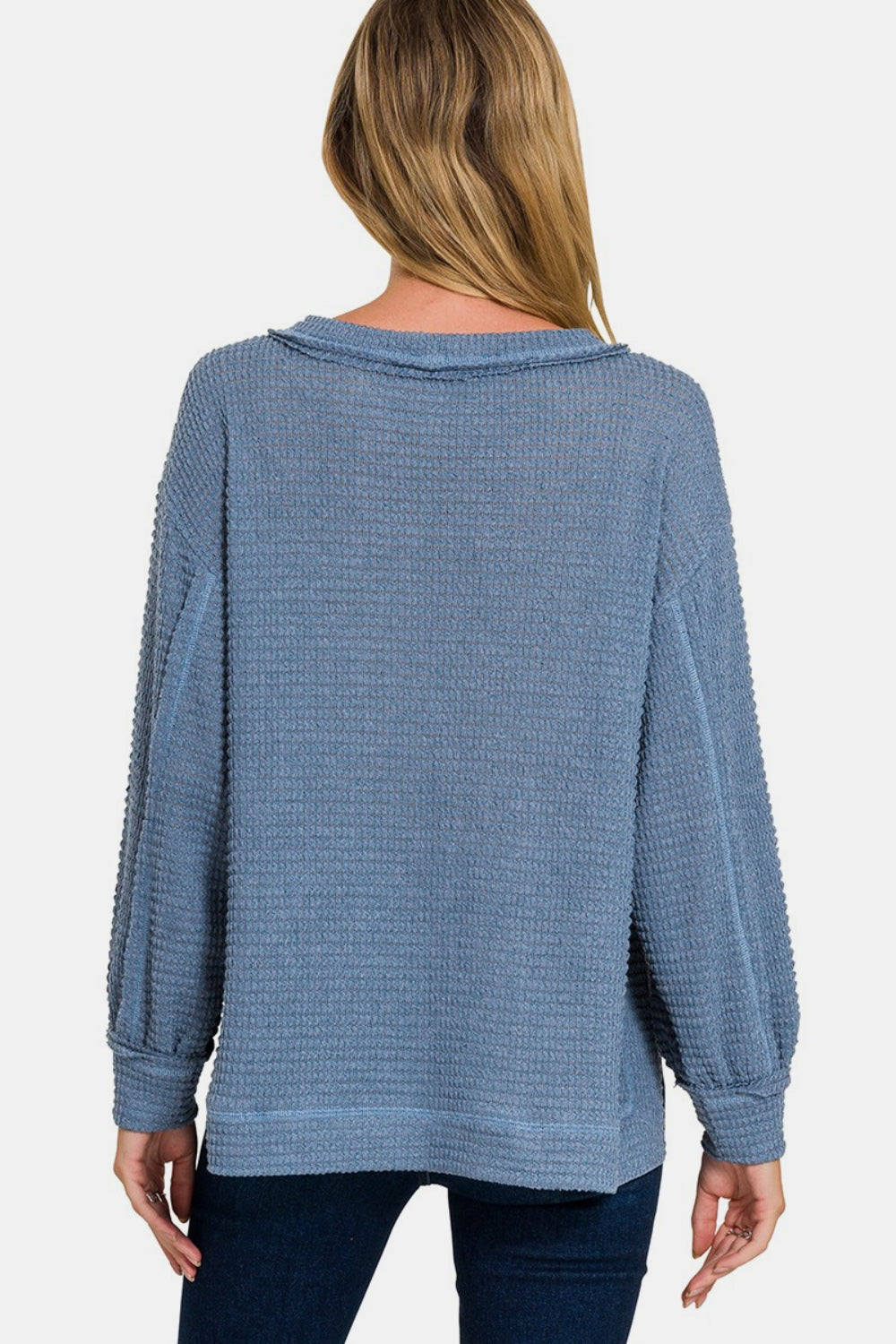 A woman wearing the Zenana Exposed Seam Side Slit Long Sleeve Top, which features a loose fit and textured blue fabric with side slits and a pocket on the left side, stands against a plain, light background. She has blonde hair and is looking directly at the camera.