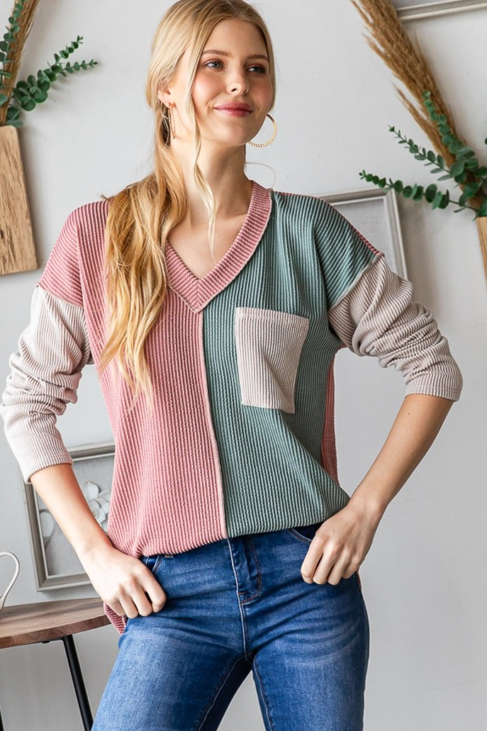 A person with long blonde hair stands indoors, smiling. They are wearing the Heimish Color Block Exposed Seam Ribbed T-Shirt, featuring pink, green, and beige sections, paired with blue jeans. Decorative wall pieces hang in the background.