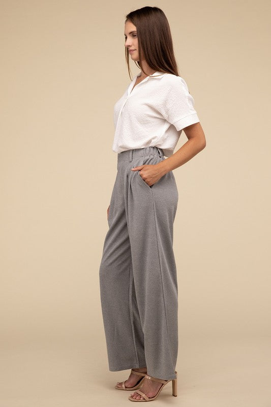 A woman stands against a beige background, highlighting her chic ensemble with a tailored fit. She wears a white short-sleeve shirt paired with versatile pink Waffle Trouser Pants, hands casually in pockets, complemented by stylish strappy sandals.