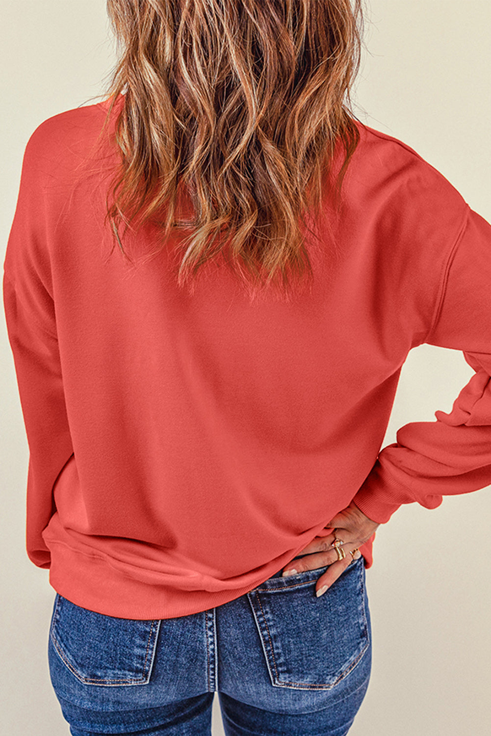 A person with wavy brown hair, wearing a Plain Crew Neck Pullover Sweatshirt in red and blue jeans, is facing away from the camera with their left hand placed on their hip.