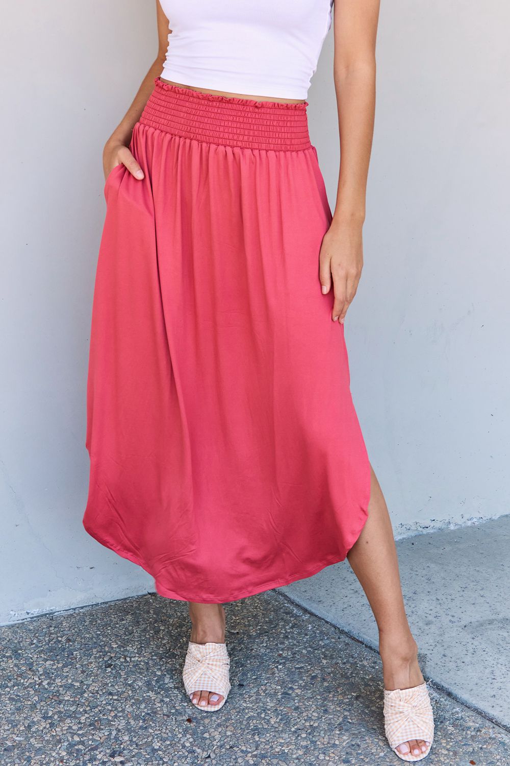 A person wearing a white top and the Doublju Comfort Princess Full Size High Waist Scoop Hem Maxi Skirt in an elegant coral color, paired with tan sandals, stands in front of a plain wall.