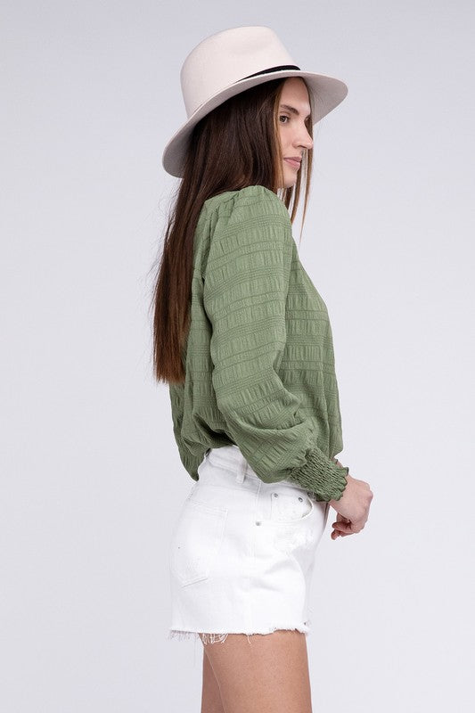 A woman wearing a Textured Shirred Yoke Top with bishop sleeves, complemented by a white hat and white shorts, stands against a plain background.