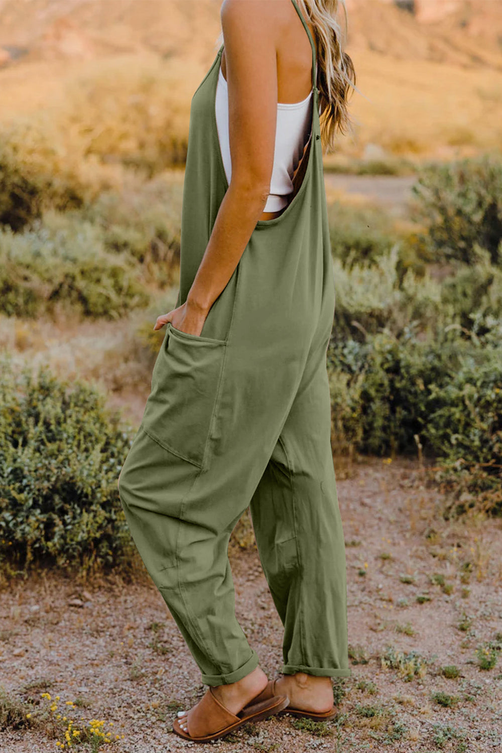 Person wearing a Double Take Full Size V-Neck Sleeveless Jumpsuit with Pockets and sandals, walking outdoors on a dirt trail with bushes in the background.