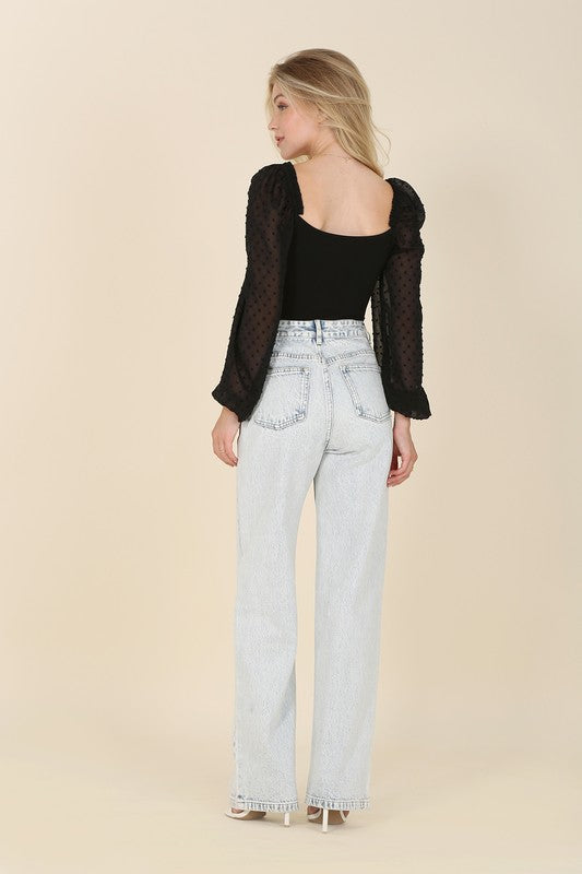 A person wearing the LS shirring sleeve bodysuit with Swiss dot mesh, featuring sheer, long puffed sleeves and a sweetheart neck, paired with light-colored jeans, stands against a plain background.