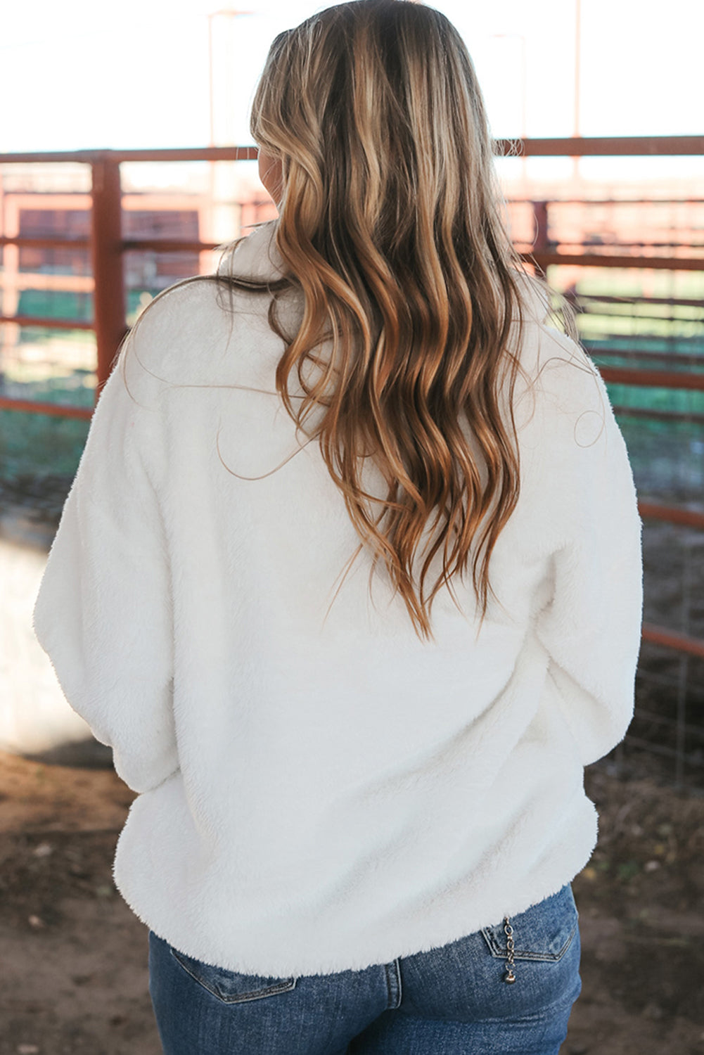 A woman with long, wavy blonde hair is seen from behind, wearing a White Leopard Patch Pocket Half Zipped Fleece Sweatshirt and blue jeans while standing outdoors near red metal fencing.