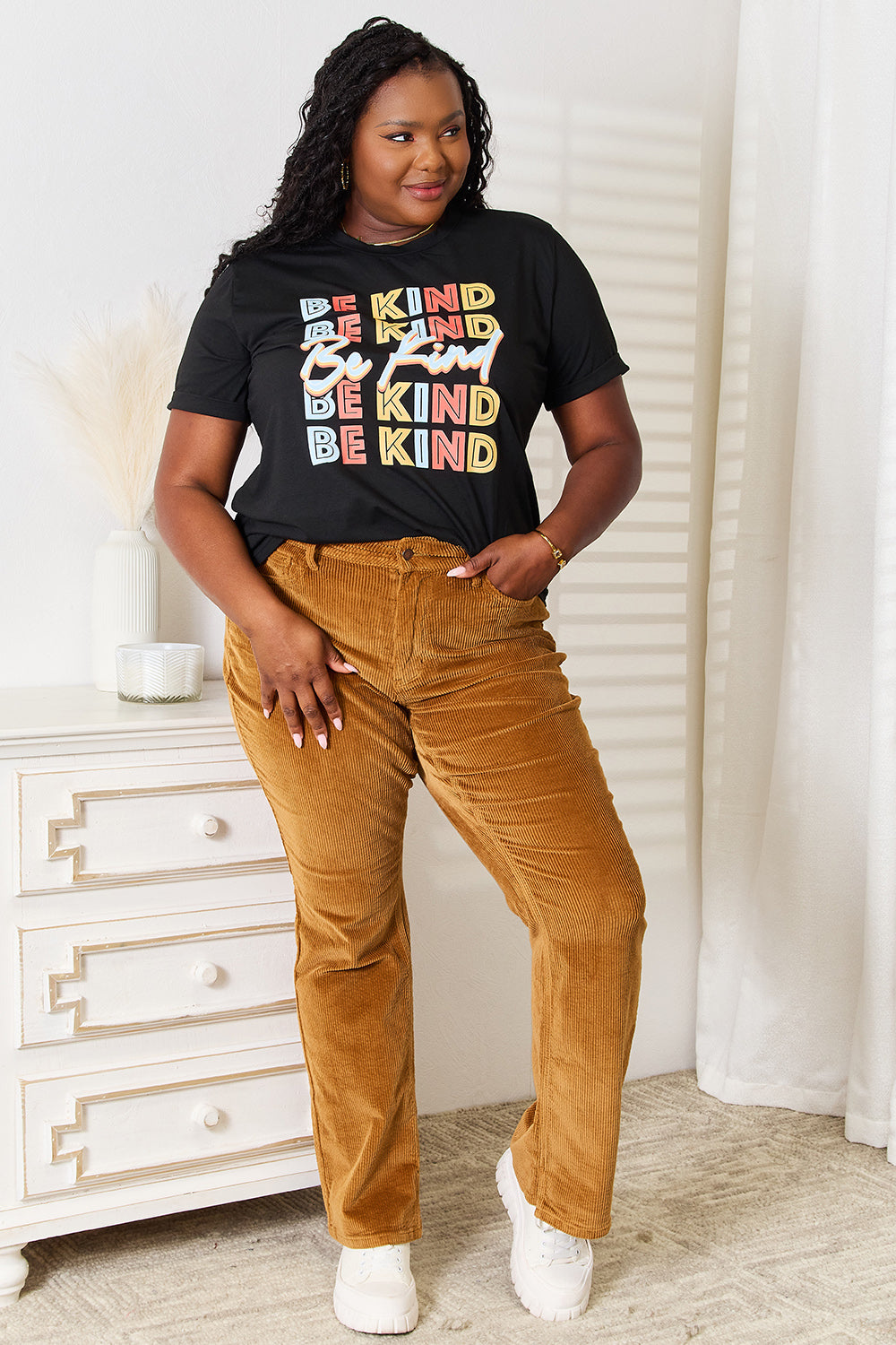 A woman stands with her hands in the pockets of her brown corduroy pants, wearing a casual Simply Love BE KIND Graphic Round Neck T-Shirt that reads "BE KIND" in large colorful letters, showcasing positive message clothing.