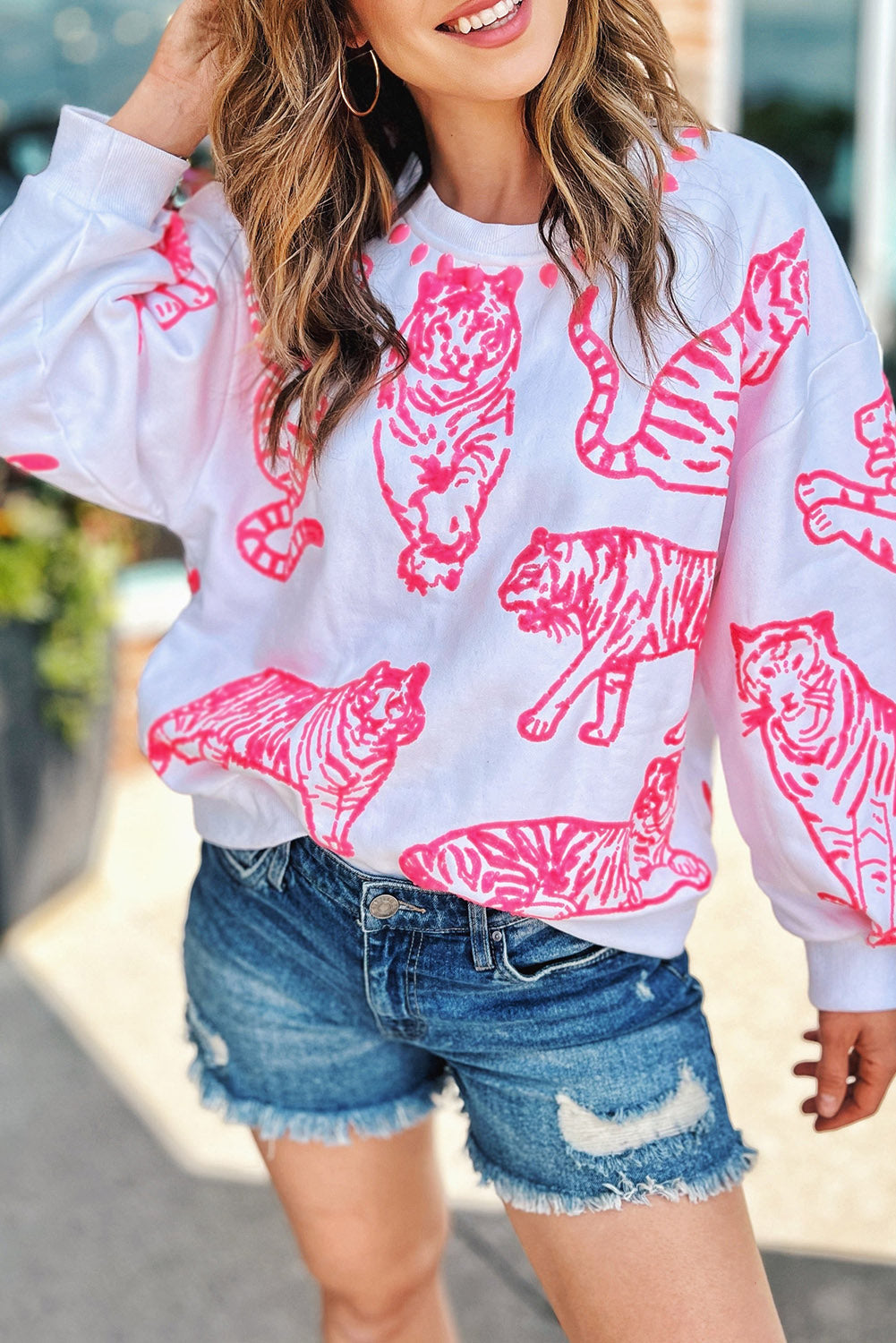 A person with long blonde hair wears a trendy Bright White Pink Animal Print Pullover Sweatshirt, paired with blue jeans, standing outdoors with their back facing the camera.