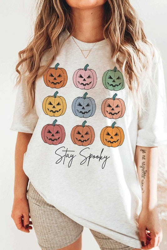 A person wears the STAY SPOOKY MULTI PUMPKINS Graphic Tee in unisex sizing, showcasing nine colorful jack-o'-lanterns and "Stay Spooky" text in a white hue, while holding a drink. They also sport checkered shorts.