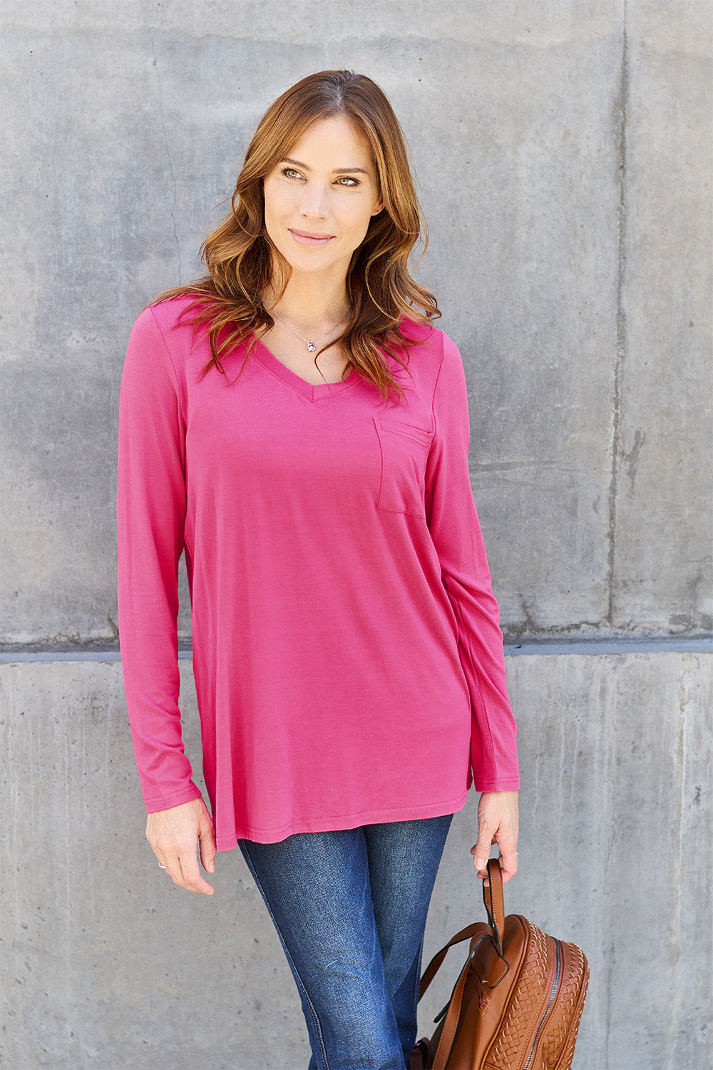 A woman with light brown hair stands against a concrete wall, wearing the Basic Bae Full Size V-Neck Long Sleeve Top in olive green and blue jeans, with a white backpack on her shoulder. Her outfit exudes basic style and is made from polyester, ensuring it’s both comfortable and suitable for machine wash cold. She is looking at the camera.