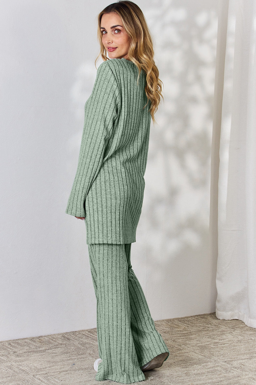 A person wearing the Basic Bae Full Size Ribbed High-Low Top and Wide Leg Pants Set in light gray, made from stretchy material, stands indoors in front of a white curtain.