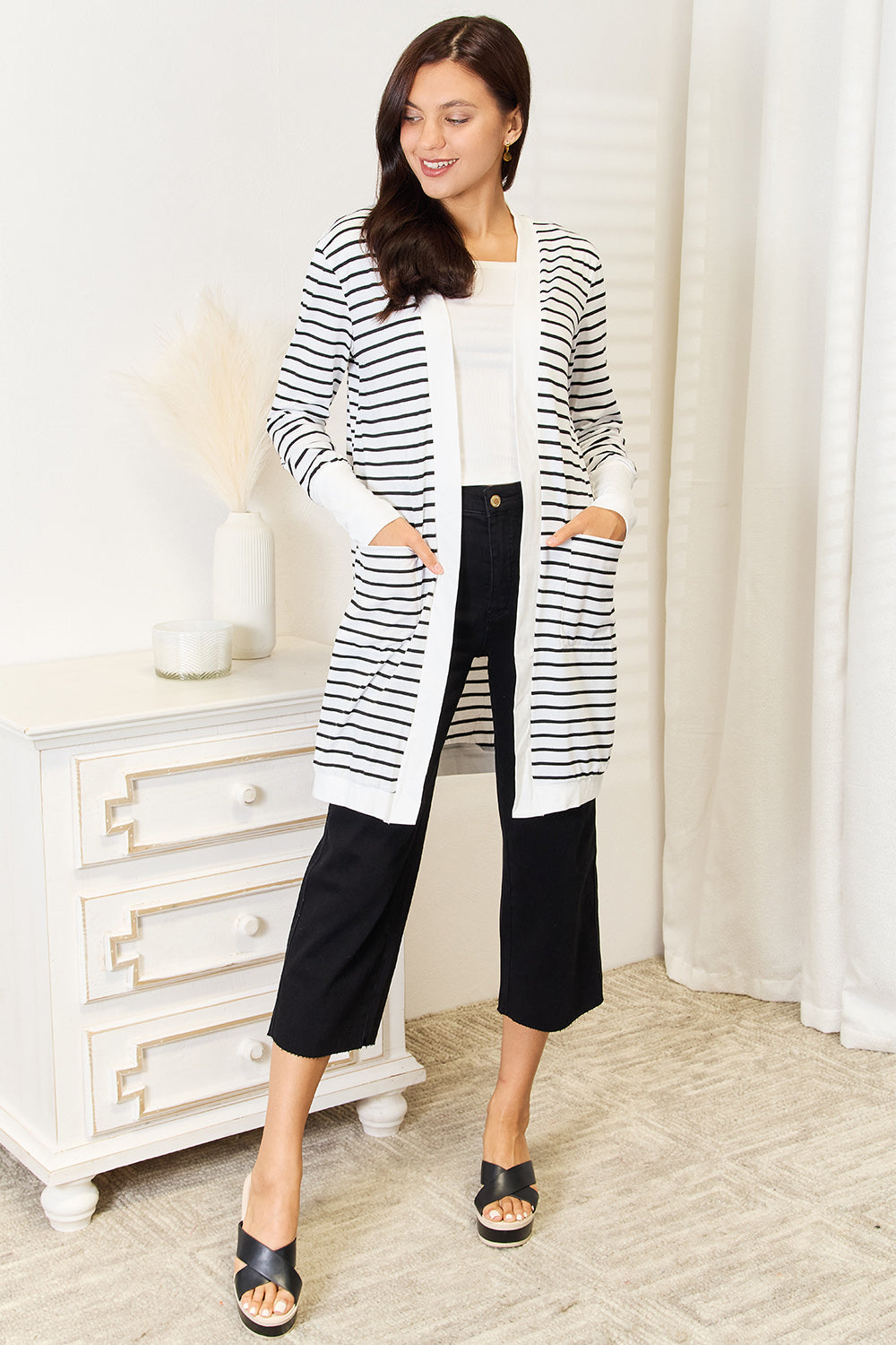 A woman wearing the Double Take Striped Open Front Longline Cardigan over a white top and black pants stands indoors, smiling at the camera. She is posing with one hand on her hip, showcasing this versatile piece.