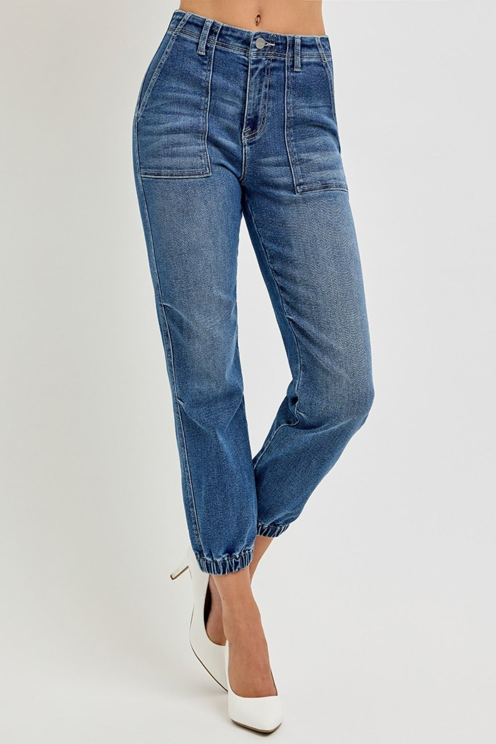 A person is wearing RISEN Full Size High Rise Ankle Jogger Jeans in light blue stretchy denim with elastic cuffs and white pointed-toe heels, standing against a plain background.