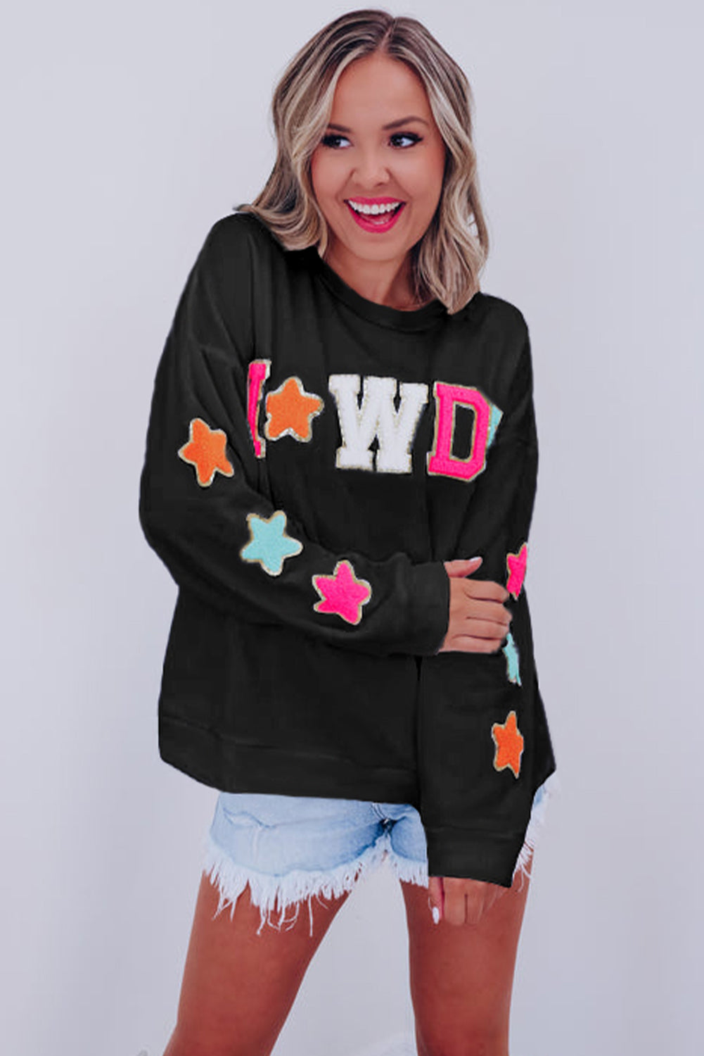 A woman with short blonde hair is wearing the Black Howdy Glitter Chenille Patch Graphic Casual Sweatshirt, featuring colorful star patterns and "WD" letters. She pairs it with light denim shorts and smiles while looking to the side.