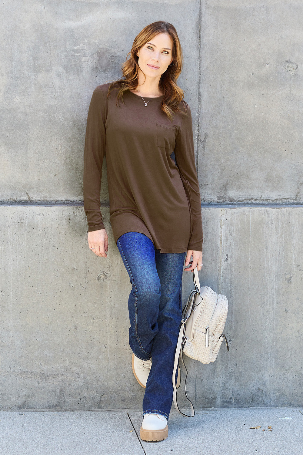 A woman with long hair wearing sunglasses, a Basic Bae Full Size Round Neck Long Sleeve Top in turquoise, and jeans is standing in front of a concrete wall with one hand on her hip.