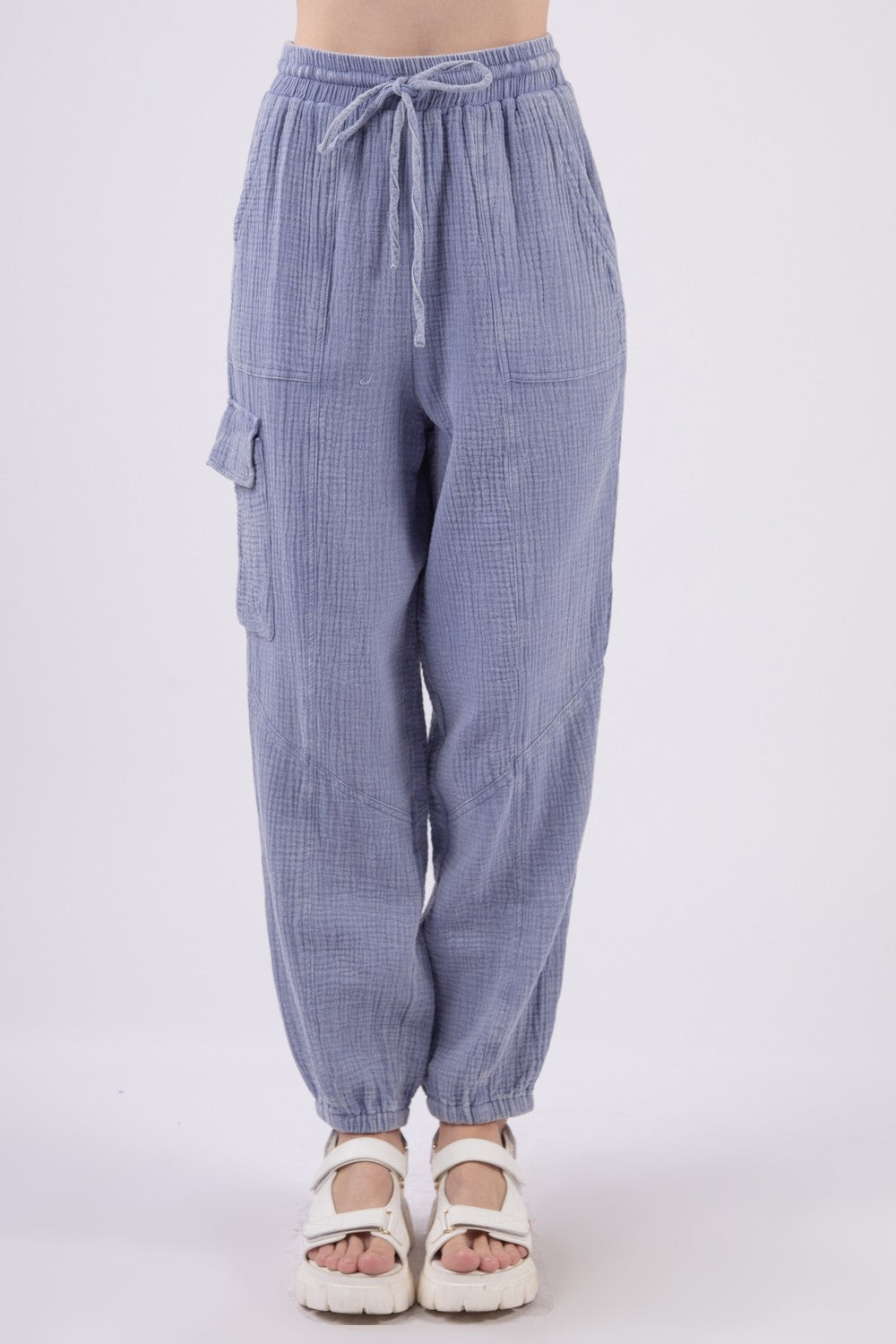 A person wearing VERY J Washed Woven Crinkle Gauze Drawstring Cargo Pants in light blue, paired with white sandals. These relaxed-fit, textured pants feature an elastic waistband and pockets, showcasing a utility design that's perfect for blending comfort and functionality against a plain background.