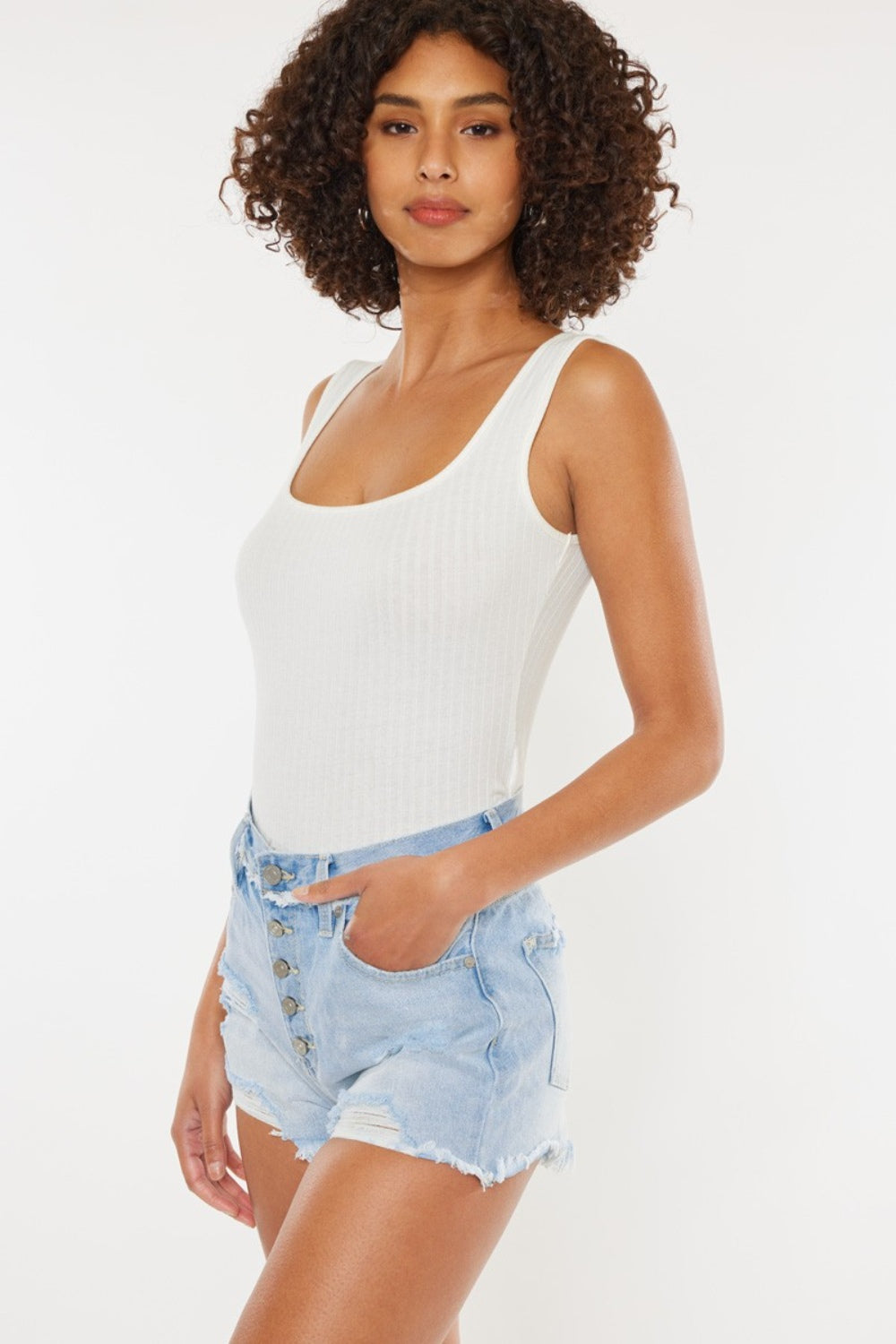 A person wearing a white tank top and Kancan Distressed Button Fly Denim Shorts poses against a plain background, showcasing a quintessential summer wardrobe staple.