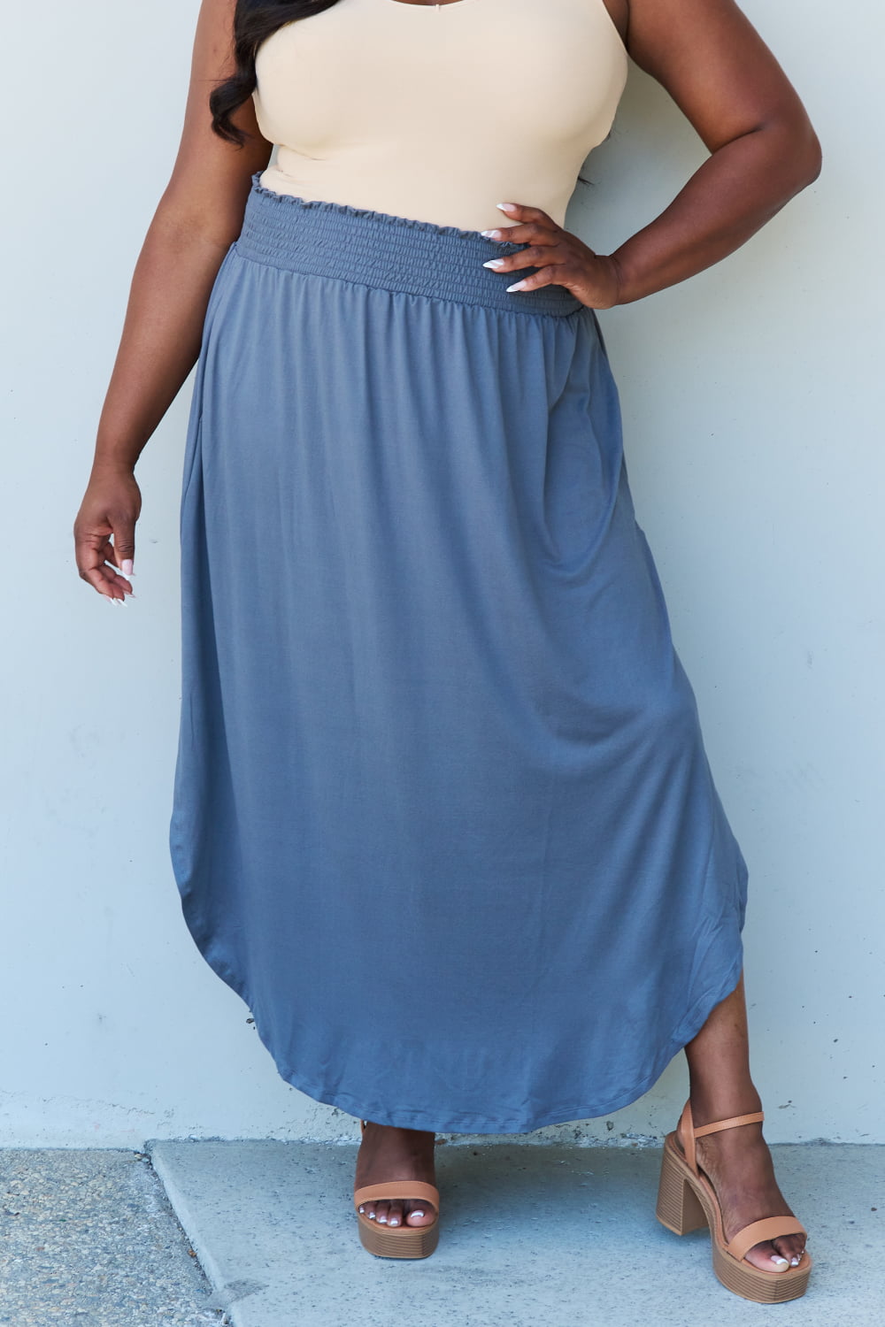 A person wearing a white sleeveless top and the Doublju Comfort Princess Full Size High Waist Scoop Hem Maxi Skirt in Charcoal stands against a plain wall. They are also wearing black sandals.