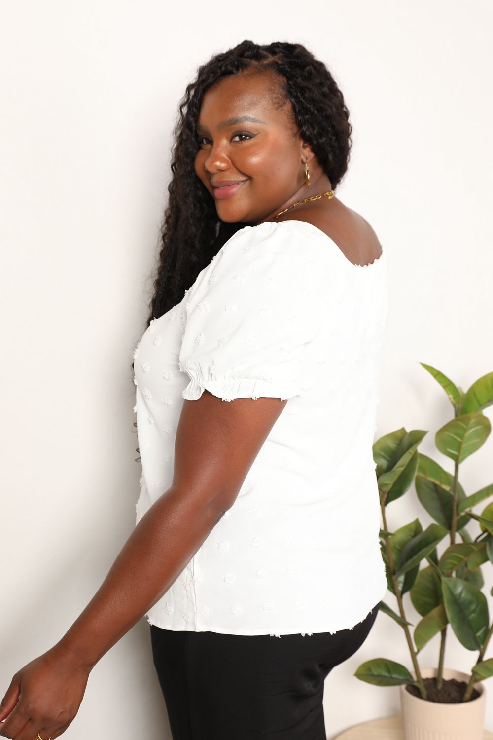 A person wearing the Mandy Swiss Dot Puff Sleeve Square Neck Blouse in white, paired with black pants, stands in front of a light background with green leaves visible on the right side.